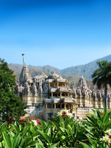 A white marble temple with intricate carvings stands amid lush greenery, inviting the adventurous traveler to explore its serene beauty, with mountainous terrain in the background under a clear blue sky.