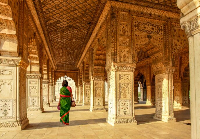 A person in a green sari walks down a richly decorated corridor with intricately carved pillars and arches, as if embarking on a timeless tour through history.