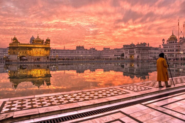 A traveler in a yellow robe stands by the water's edge, facing the illuminated Golden Temple complex at sunset, with a colorful sky reflected in the water.