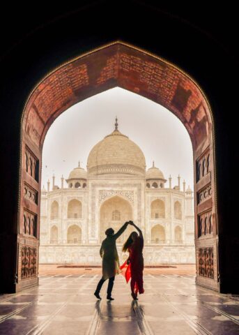 Silhouettes of a couple dance under an ornate archway in India, the majestic Taj Mahal illuminated in the background.