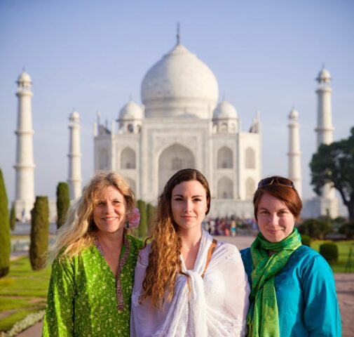 Three adventurous women stand in front of the Taj Mahal on a sunny day, with the iconic white mausoleum and its gardens clearly visible in the background.
