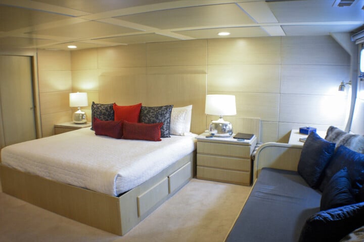 A bedroom with a large bed adorned with red and grey pillows, beige walls, two bedside tables with lamps, and a sofa against the wall invites you to dream of travel.