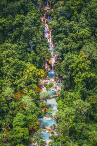 Aerial view of a multi-tiered waterfall in Laos, cascading through dense lush green forest and pooling in blue turquoise natural basins.