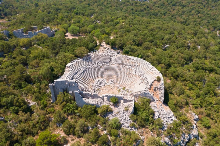 Aerial view of the ruins of an ancient amphitheater in Turkey, surrounded by a dense forest. The stone structure is circular with multiple levels of seating, partially covered by vegetation.