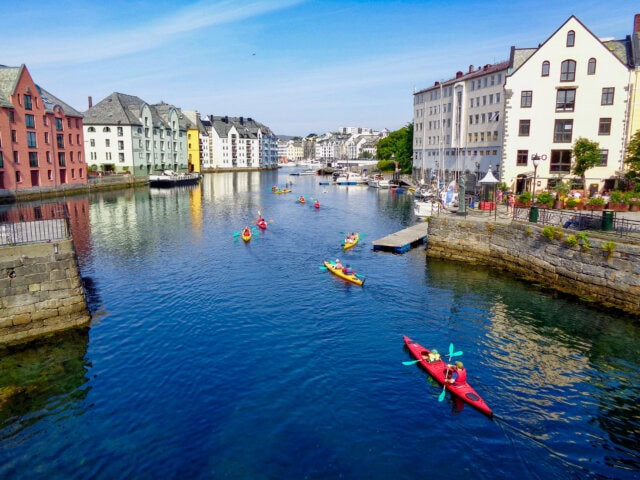 Kayakers paddle along a calm waterway lined with colorful buildings under a clear blue sky, showcasing the scenic beauty of Norway—an idyllic destination for travel and tourism.