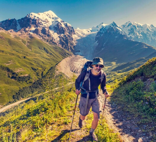 A person hiking on a narrow mountain trail in Georgia, with snow-capped peaks and a glacier in the background, under a clear blue sky.