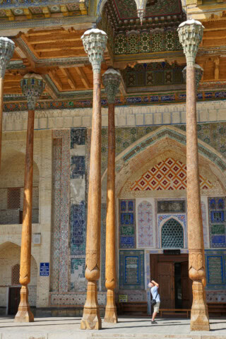 A person with a backpack enters a building in Uzbekistan, adorned with intricate colorful tilework, tall wooden columns, and detailed archways.