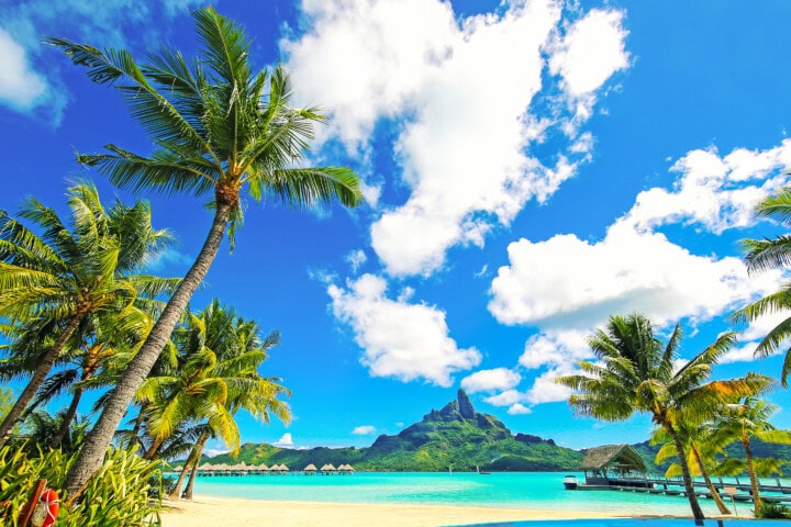Tropical beach scene with palm trees, blue ocean water, overwater bungalows, and a mountainous island in the background under a blue sky with scattered clouds—quintessential French Polynesia.