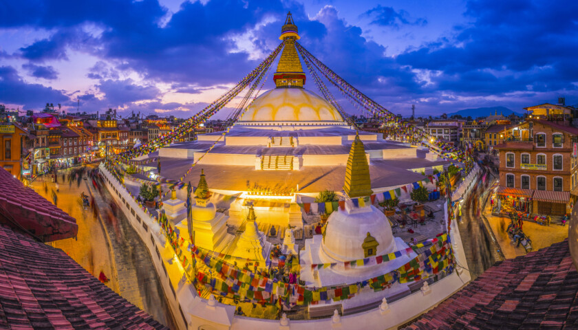 A large stupa decorated with colorful prayer flags in Nepal, surrounded by a bustling square and buildings, under a vibrant evening sky with clouds at twilight.