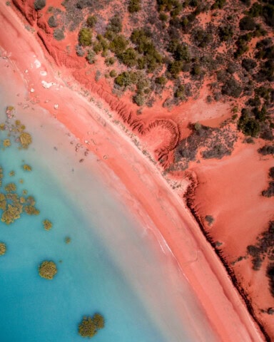 Aerial view of Australia's coastal landscape showcases a vibrant contrast between turquoise ocean water and red sandy terrain, dotted with green vegetation.