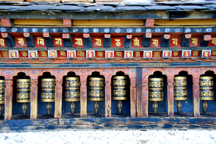 A row of Tibetan Buddhist prayer wheels featuring intricate carvings and inscriptions, set beneath a colorful wooden structure, offers a glimpse into the rich cultural tapestry that makes Bhutan an alluring travel destination.