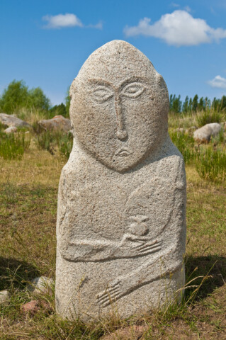A stone statue featuring a humanoid figure with minimalist facial features and crossed arms sits on a grassy field in Kyrgyzstan, with a blue sky and scattered clouds in the background.