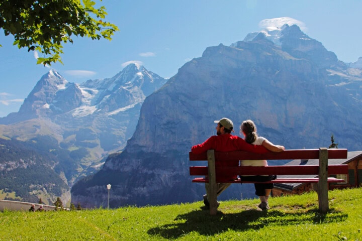 Two people sit together on a bench, facing the large, snow-capped mountains under a clear blue sky, epitomizing the serene beauty of Swiss tourism.
