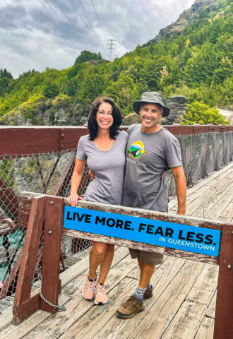 Two people standing on a wooden bridge in front of a sign that reads "Live More. Fear Less. In Queenstown, New Zealand." The background features lush greenery and power lines.