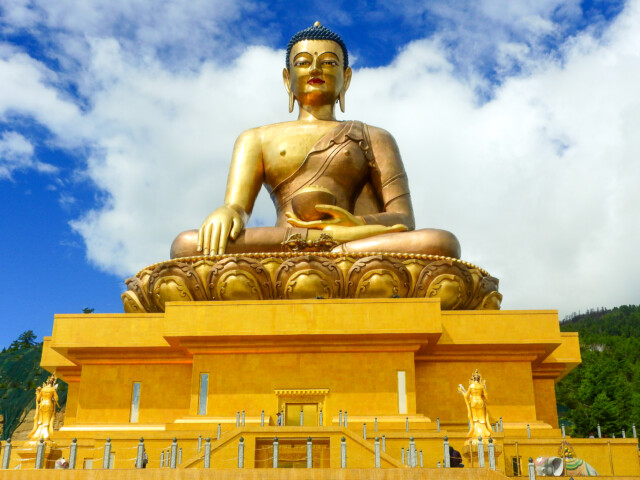 Golden statue of a seated Buddha on a large lotus pedestal against a blue sky with clouds, resembling the serene architecture found in Bhutan, surrounded by smaller statues and a railing at the base.