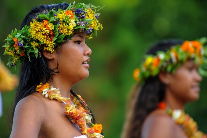 Two women wearing floral crowns and leis participate in a cultural event outdoors on the Polynesian islands. The woman in the foreground gazes to the side, while the background is lush and green.