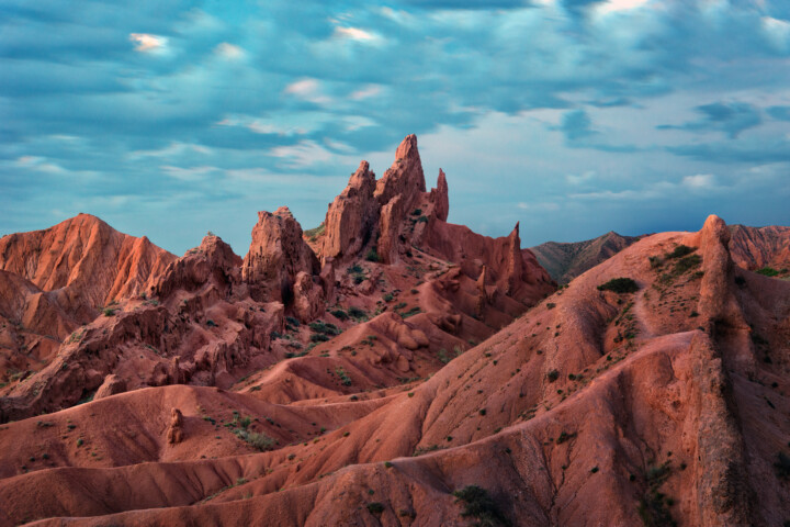 Jagged red rock formations rise sharply against a backdrop of a cloudy sky and the rugged landscape of Kyrgyzstan.