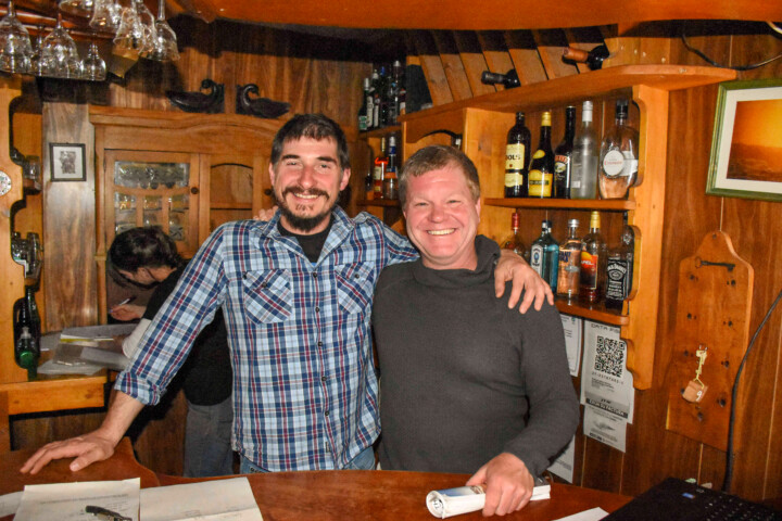 Two men, both smiling, stand behind a wooden bar adorned with various bottles. One man has his arm around the other, exuding camaraderie. In the background, another person is busy writing. The scene evokes the warm hospitality often found in Argentina's vibrant bars.