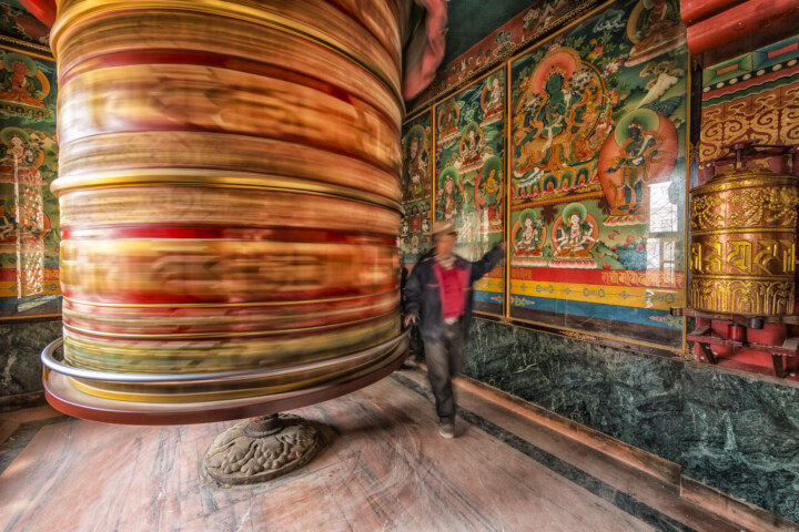 A person spins a large painted prayer wheel inside a richly adorned room in Nepal, with colorful murals depicting spiritual figures.