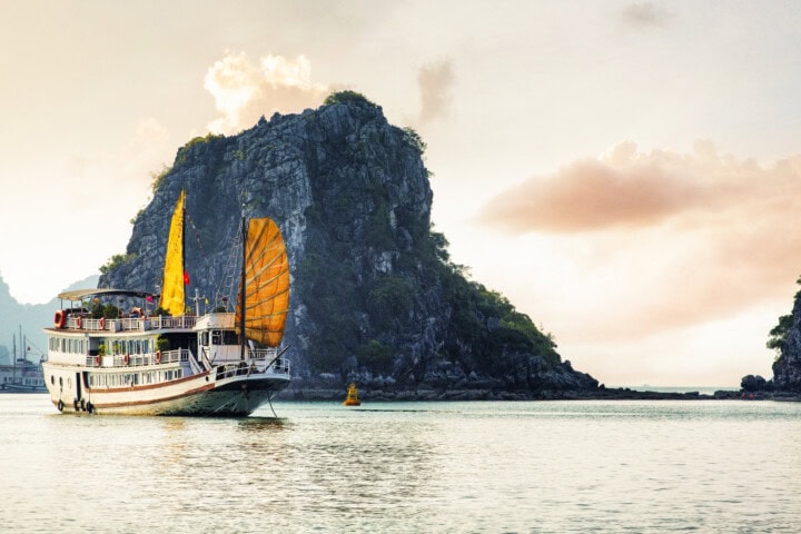 A boat with yellow sails is on the water near a rocky, forested island under a cloudy sky at sunset, evoking the serene beauty of Vietnam's coastal landscapes.