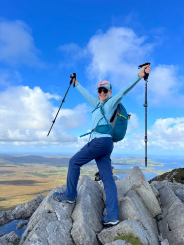 A person wearing outdoor gear and sunglasses stands triumphantly on rocky terrain, holding hiking poles with both arms outstretched against a backdrop of open skies and the distant landscapes of Ireland, encapsulating the spirit of adventure in travel tourism.