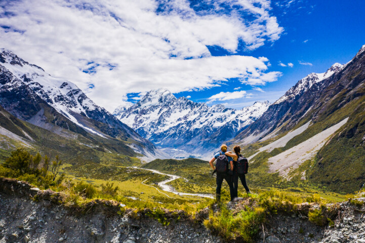Two hikers with backpacks stand on a rocky ridge, overlooking a scenic valley with snow-capped mountains under a partly cloudy sky in New Zealand.