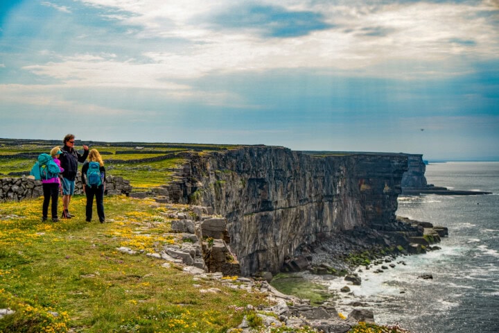 Four people stand on a grassy clifftop in Ireland, overlooking a rugged coastline with cliffs and the sea under a partly cloudy sky.