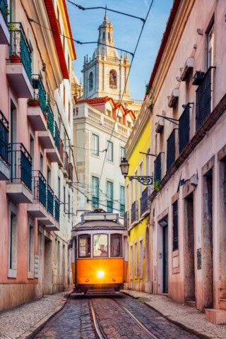A yellow tram travels along a narrow street lined with colorful buildings, offering a quintessential slice of Portugal's charm. In the background, under a clear sky, stands a church tower—a perfect capture for anyone interested in travel and tourism.