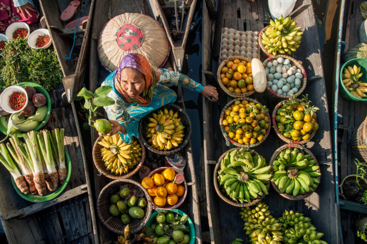 A woman in a boat at a Malaysian floating market sells various fruits and vegetables, including bananas, limes, and eggs. Fresh produce is displayed in baskets around her.