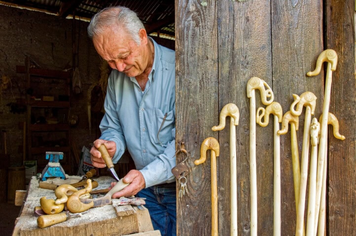 An elderly man carves a wooden cane in a workshop. Several finished canes with curved handles stand against a wooden door beside him, reminiscent of the artisanal craftsmanship seen in Greece.