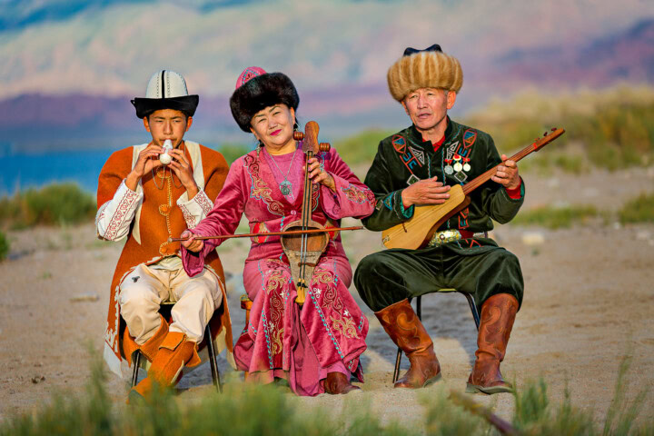 Three performers in traditional attire play musical instruments on a grassy landscape with the majestic mountains of Kyrgyzstan in the background.