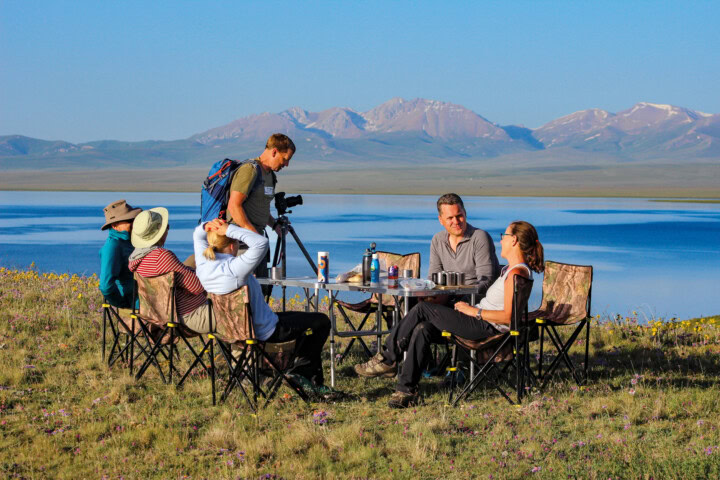Five people sit and stand around a table with a camera on a tripod in front of a lake, set against the breathtaking mountains of Kyrgyzstan. This picturesque scene captures the essence of Central Asia and highlights the unique allure of tourism in this stunning region.