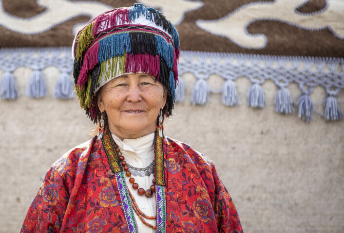 An elderly woman in traditional colorful attire and a fringed hat stands in front of a decorated wall in Kyrgyzstan, smiling.
