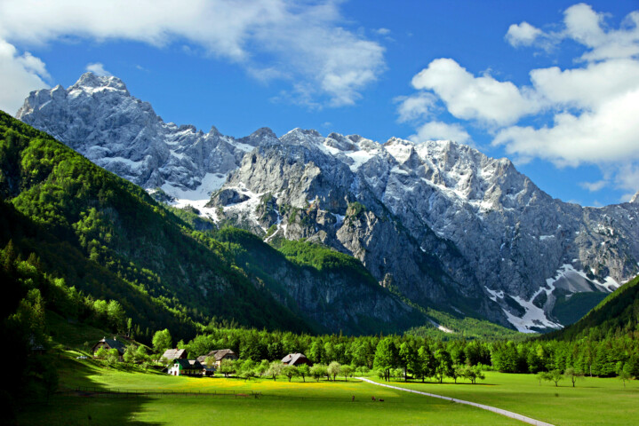 A serene landscape featuring a lush green valley, scattered houses, and a winding road, set against a backdrop of towering, snow-capped mountains under a blue sky with scattered clouds in picturesque Slovenia.