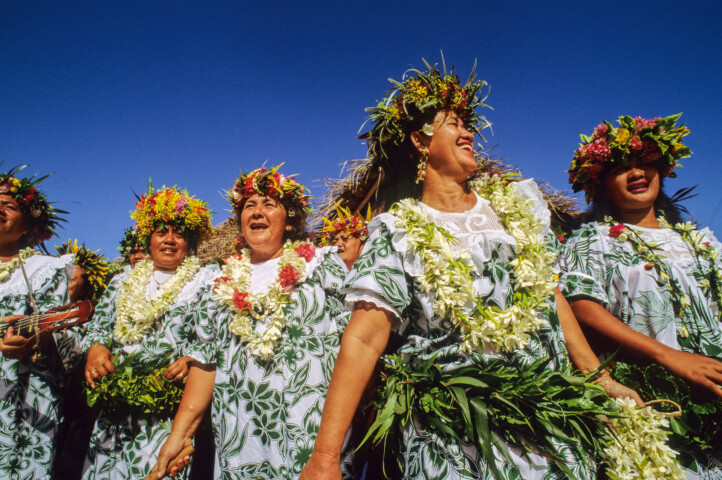 A group of people in French Polynesia, adorned with floral crowns and green and white outfits, perform a traditional dance with leis around their necks against the backdrop of a clear blue sky.