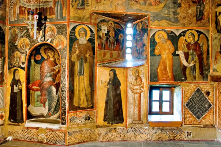 A richly decorated religious mural featuring various saintly figures and scenes, with vibrant colors and intricate designs. A small window is embedded within the mural, adding a unique charm that draws tourism from visitors eager to experience the beauty of Greece.