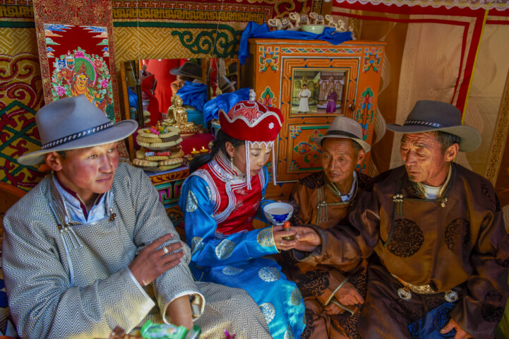 Several people in traditional Mongolian attire sit together in a decorated tent. One individual in a blue and red outfit holds a cup, while the others are engaged in conversation.