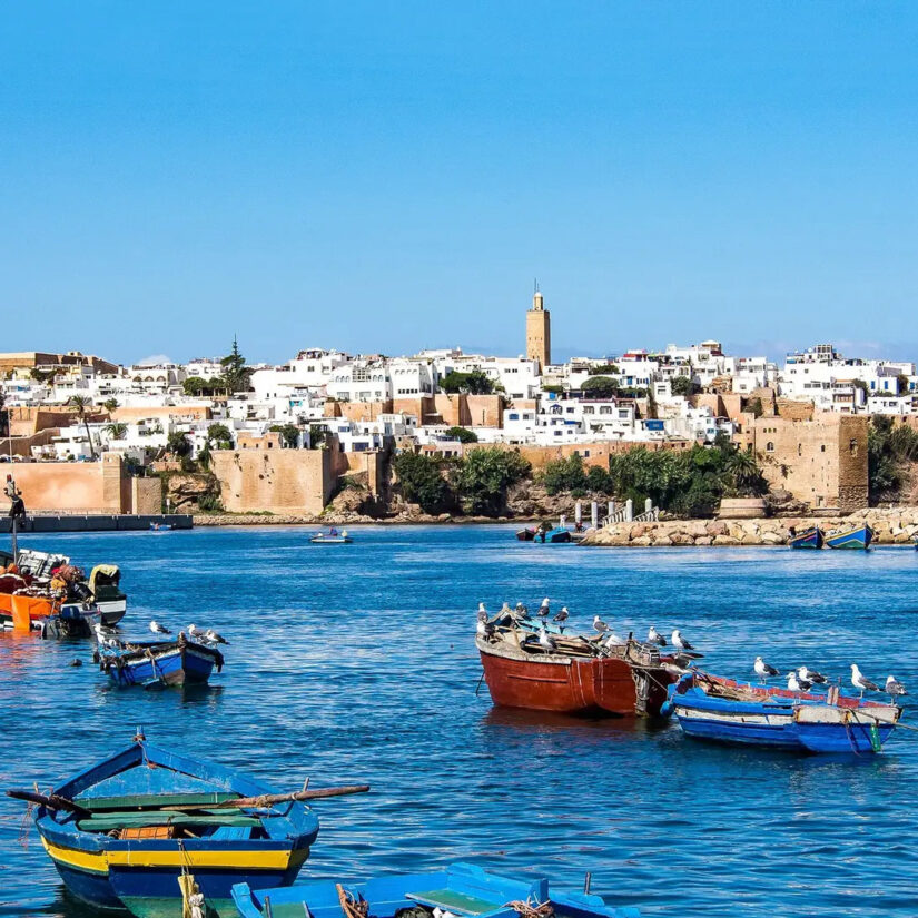 Small blue, red, and yellow boats bob in the blue water in front of a Moroccan cityfront.
