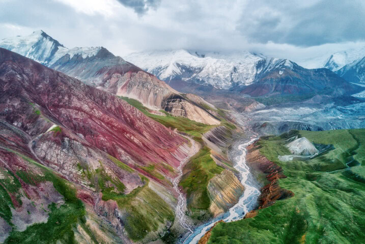 Aerial view of the stunning Kyrgyzstan mountains with red and green slopes, a winding river running through the valley, and snow-capped peaks in the background under a cloudy sky.