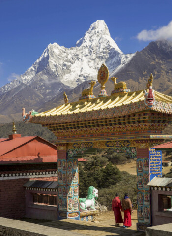 Two monks in red robes stand at an ornate gateway with intricate designs, against the backdrop of a snowy mountain peak in Nepal.