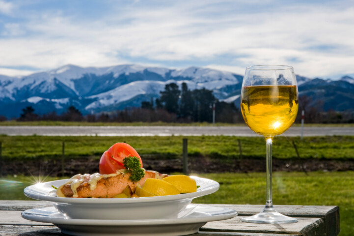 A plate of food with a lemon garnish and a glass of white wine on a wooden table, set against the breathtaking backdrop of New Zealand's snow-capped mountains and clear blue sky.