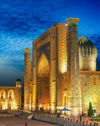Night view of a mosque in Uzbekistan with intricate geometric patterns and detailed architecture, featuring two tall minarets and a large dome, illuminated against a twilight sky.