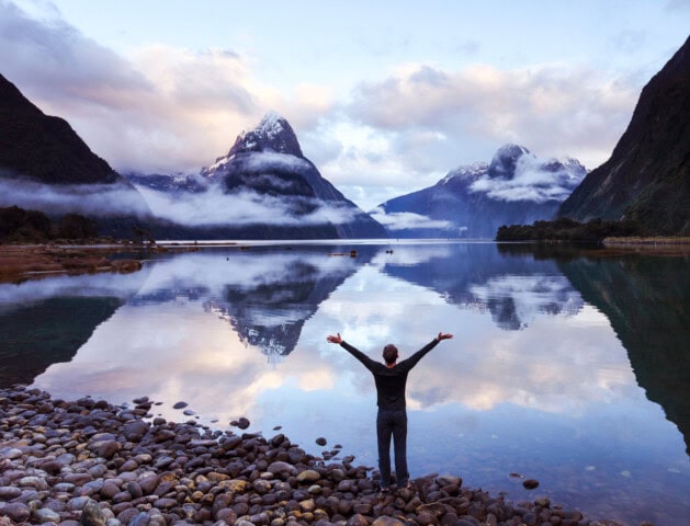 Person standing on rocky shore with arms raised, overlooking a calm lake in New Zealand reflecting snow-capped mountains and clouds.