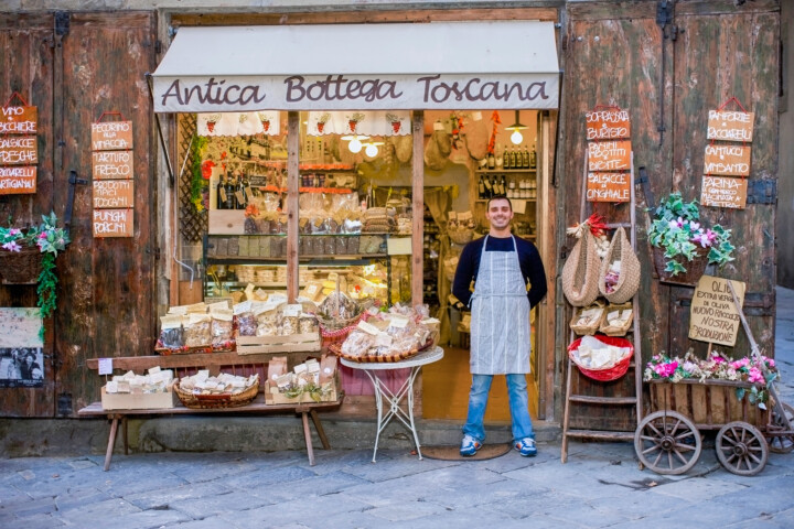 A shop owner stands in front of "Antica Bottega Toscana," a store with a rustic wooden facade, displaying a variety of traditional Italian goods and products outside, inviting travelers to experience the charm of Italy's rich heritage.