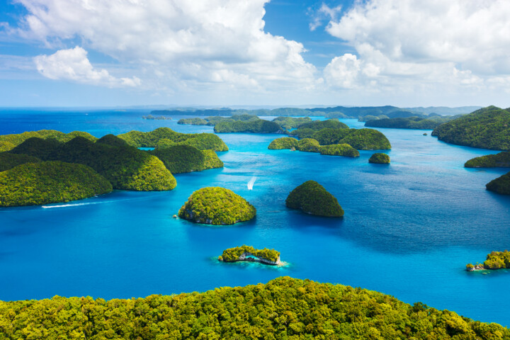 Aerial view of numerous green islands surrounded by clear blue ocean water under a partly cloudy sky, capturing the breathtaking beauty of Palau—a must-visit destination for travel and tourism enthusiasts.