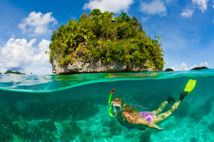 A person snorkeling in clear blue water near a small, lush island under a bright sky with scattered clouds in Palau.