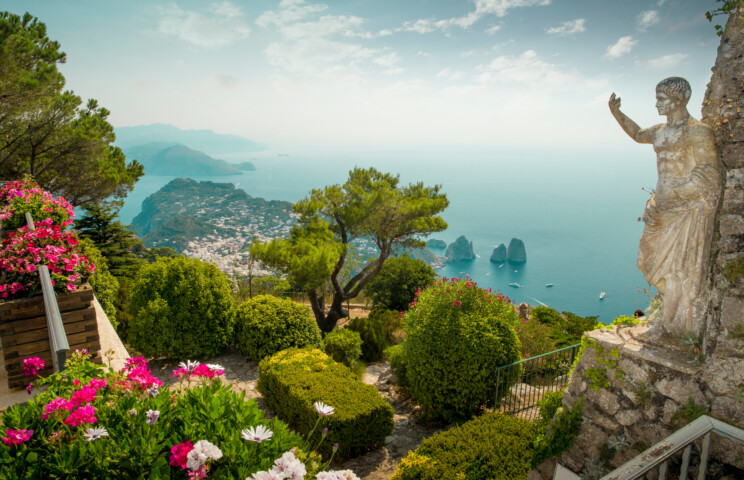 A scenic view of Italy's coastal landscape with lush greenery, vibrant flowers, a stone statue, and rocky islands in the blue sea under a clear sky.