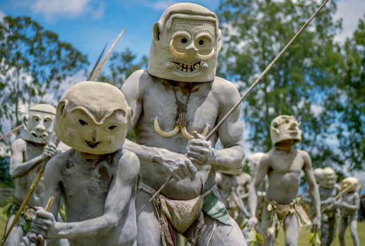 People wearing traditional mud masks and body paint, holding spears, gather outdoors with trees in the background in Papua New Guinea.
