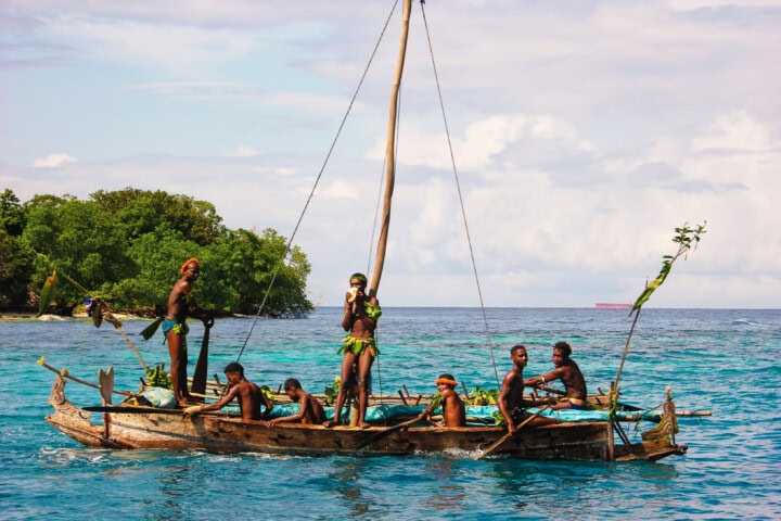 A group of people navigate a small wooden sailboat in the clear blue waters near the lush, green shoreline of Papua New Guinea.
