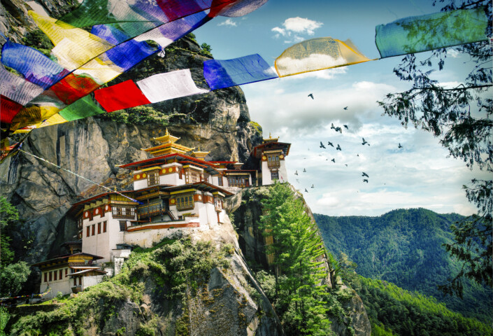 A monastery built on a rocky cliff in Bhutan is surrounded by lush mountains under a blue sky, with colorful prayer flags and flying birds in the foreground—a perfect scene for tourism and travel enthusiasts.
